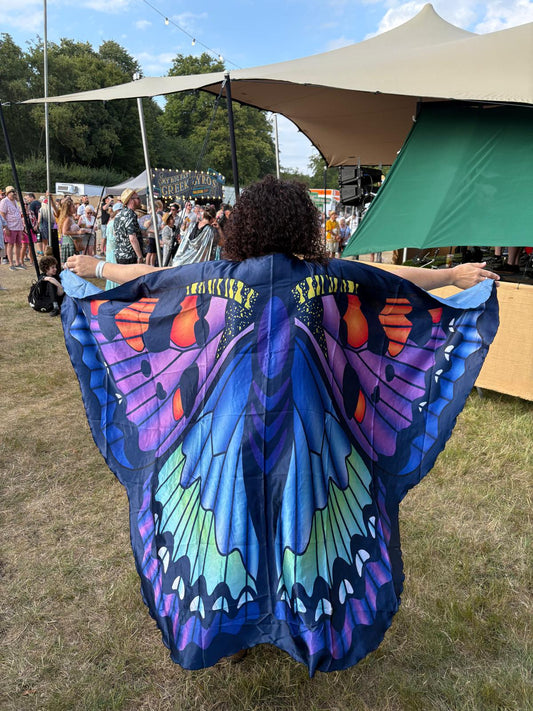 Person wearing a colorful blue butterfly costume at an outdoor event with tents and people in the background.