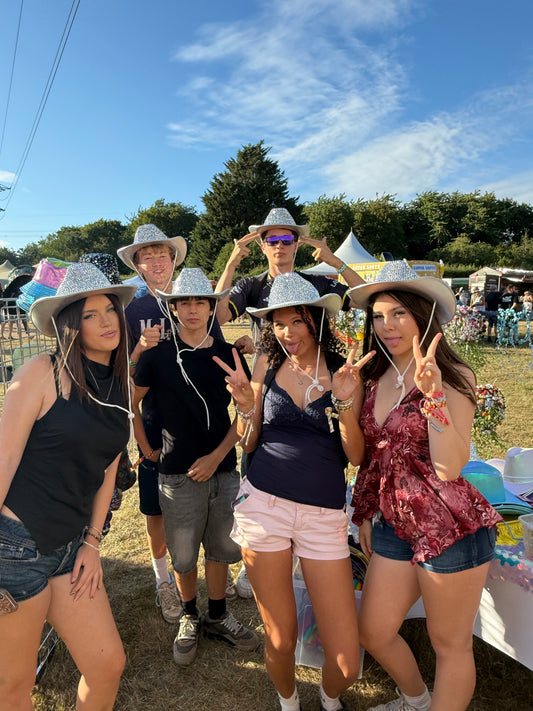 Five young women wearing matching sparkly silver cowboy hats pose together outdoors at a sunny festival or fair, making peace signs against a background of trees and blue sky.